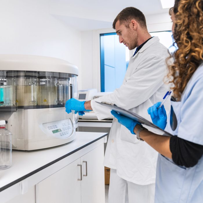 Doctor points at an analyzer while a nurse takes notes, both in lab coats and gloves in a sterile hospital lab.